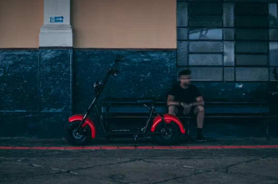 Man sitting next to red and black scooter; image by Vinicius Löw, via Unsplash.com.